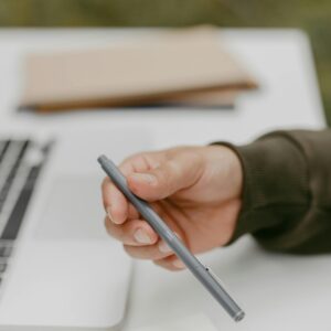 Close-up of a person holding a ballpoint pen near a laptop, symbolizing work or study.